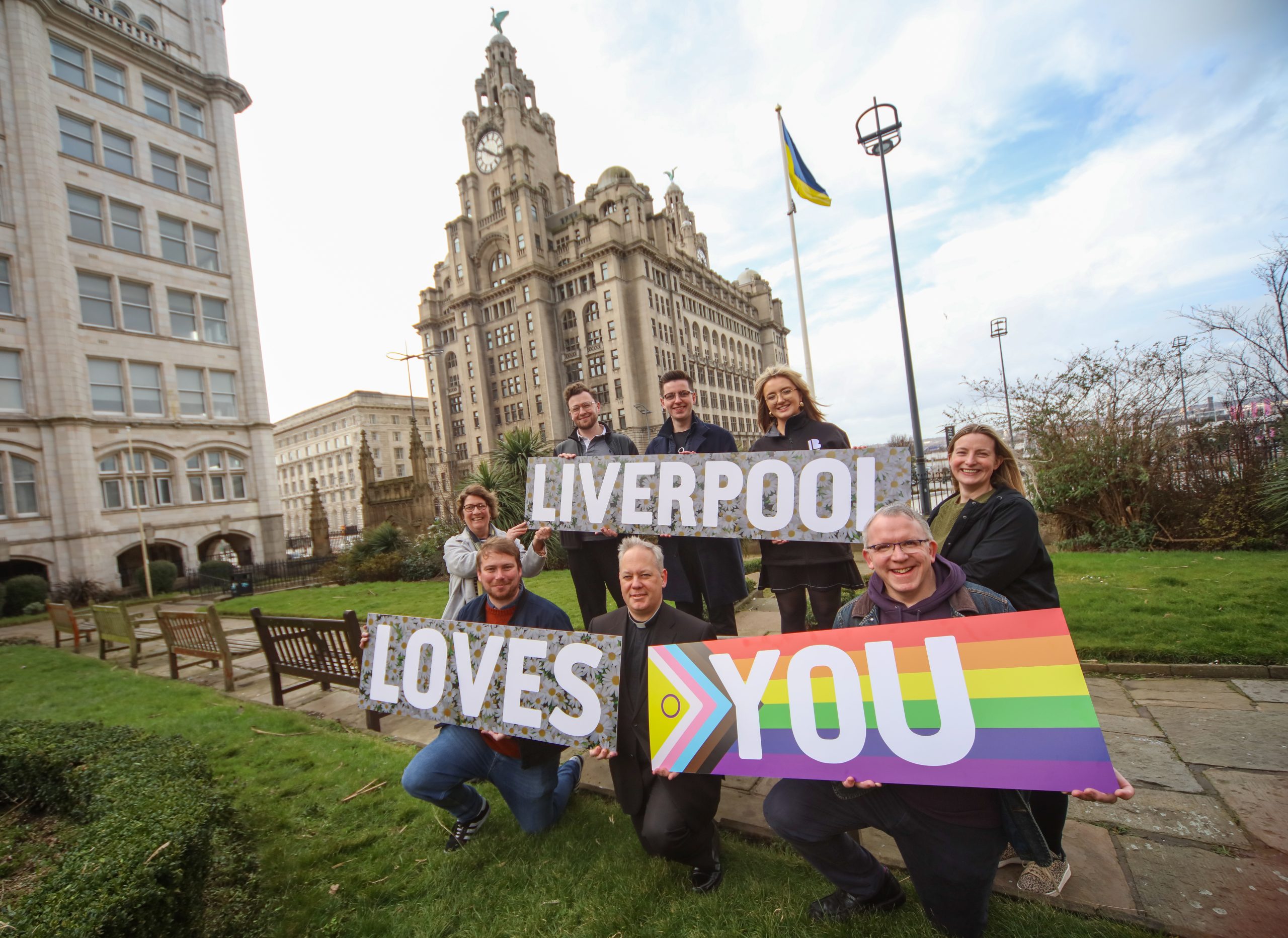 Members of the team organising the Liverpool Loves You event hold aloft a 'Liverpool Loves You' sign in St Nichols Gardens with the Liver Building in the background.