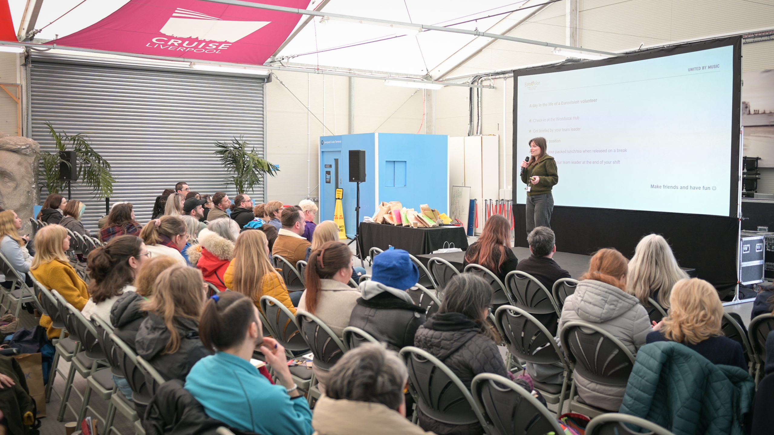 People in a room watching and listening to a person talking on a stage
