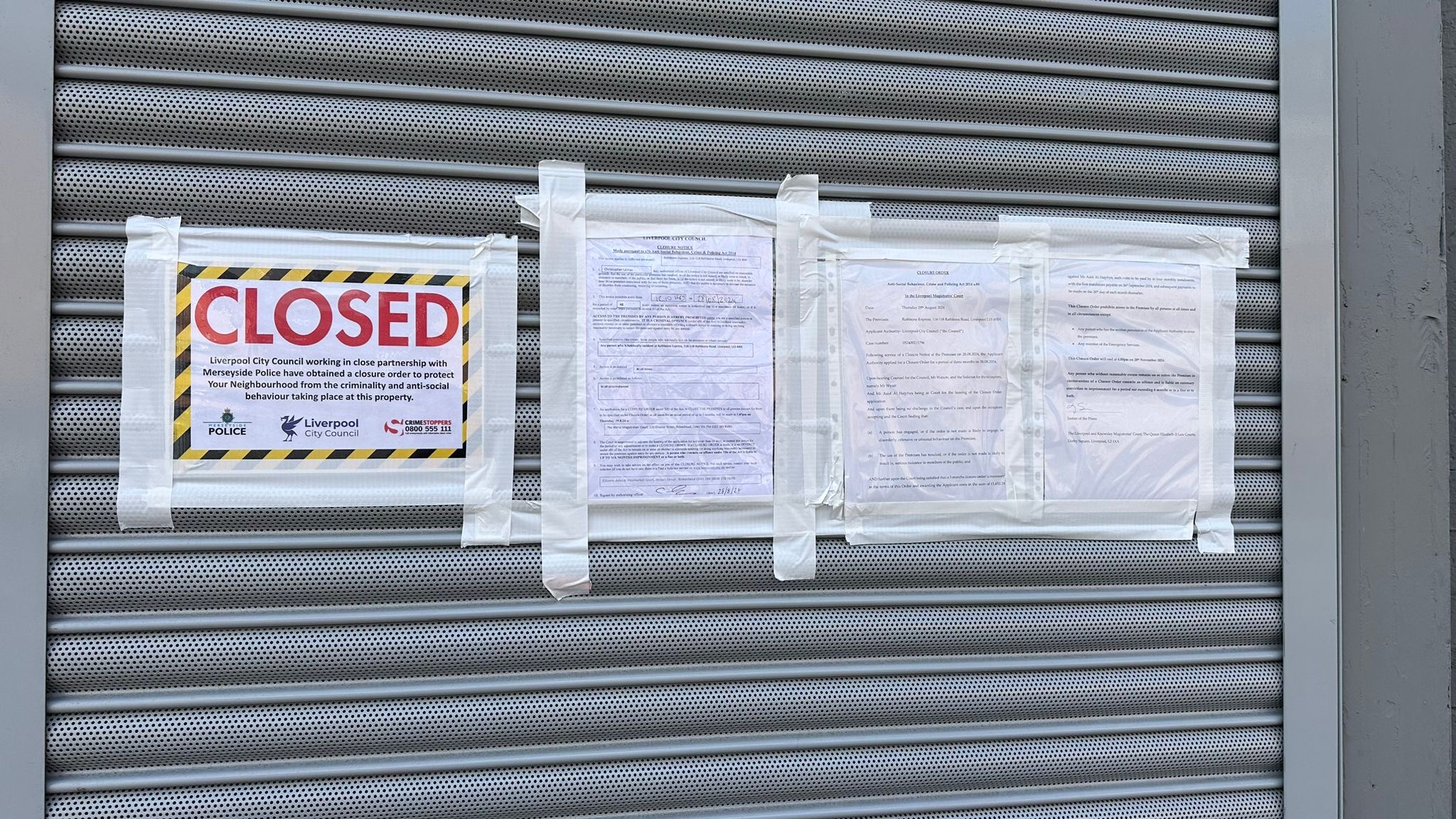 The image shows a metal roller shutter door with several notices taped to it. The most prominent notice in the centre reads "CLOSED" in large red letters. Below that, the notice states that Liverpool City Council, in close partnership with Merseyside Police, has obtained a closure order to protect the neighbourhood from criminality and anti-social behaviour taking place at this property. The notice includes the logos of Merseyside Police, Liverpool City Council, and Crimestoppers. There are additional legal documents taped next to this notice on either side, likely detailing the closure order or related legal information.