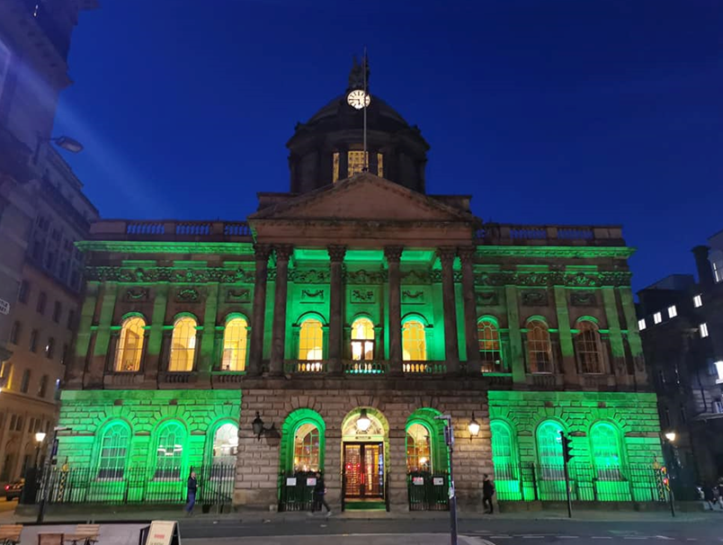 Liverpool Town Hall illuminated in green at night to mark World Mental Health Day. The historic building's classical architecture is highlighted by the green lighting, with a dark blue sky as the backdrop. Pedestrians can be seen near the entrance.