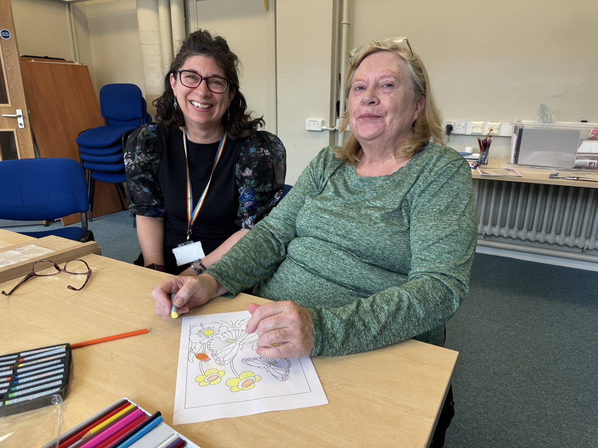 two woman sitting together smiling. One of the woman is colouring in
